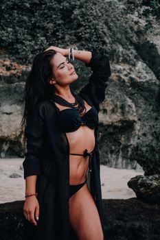 Woman in black bikini posing on a rocky beach with eyes closed, embracing summer vibes.