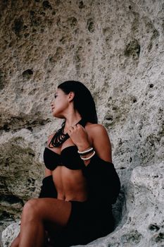 A fashionable woman in a black bikini top poses confidently against a rocky background.