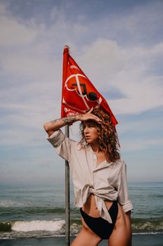 A fashionable woman poses on a beach with a red flag under a clear sky.