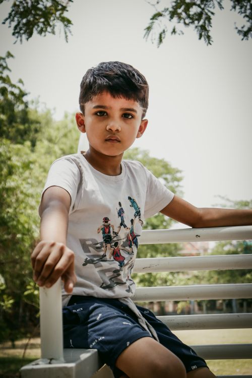 Free Young boy sitting on a bench holding railing in a sunny park. Stock Photo