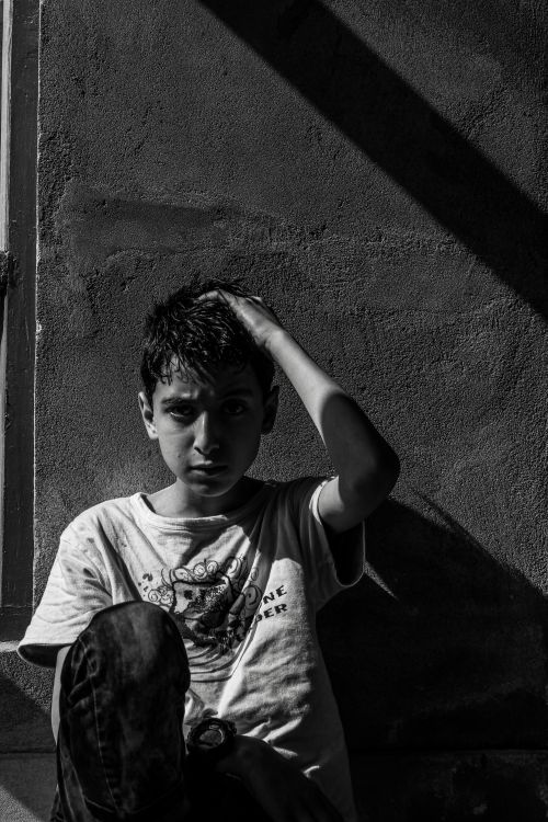 Free Black and white portrait of a boy sitting against a wall, hand on head, thoughtful expression. Stock Photo