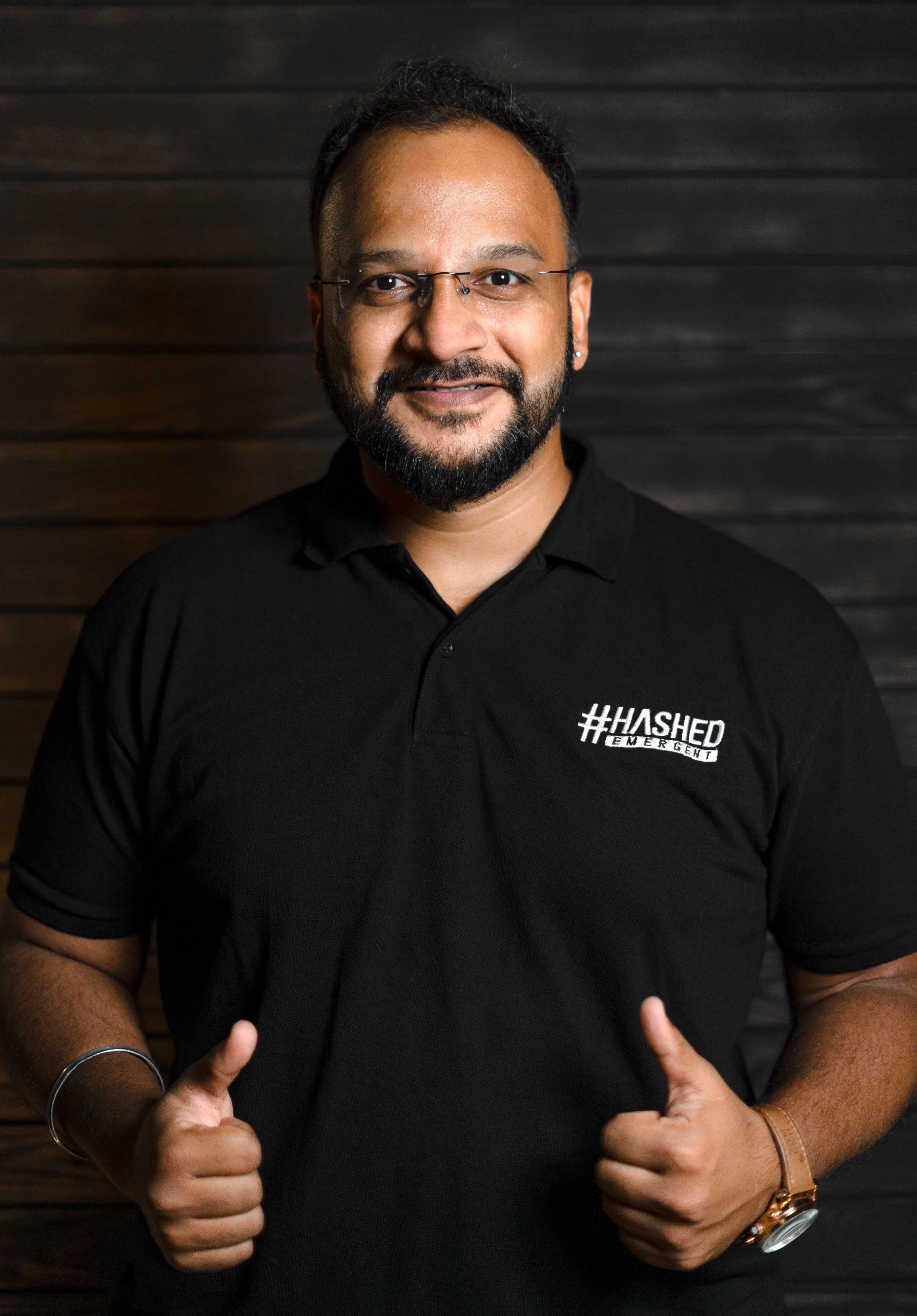 Portrait of a smiling man in a black polo shirt giving thumbs up against a wooden background.