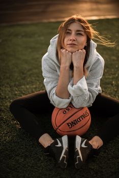 Woman in gray hoodie posing with basketball on outdoor field during sunset.