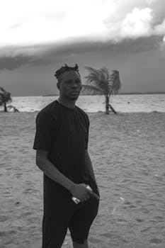 Black and white photo of a man standing on a sandy beach with a cloudy sky.