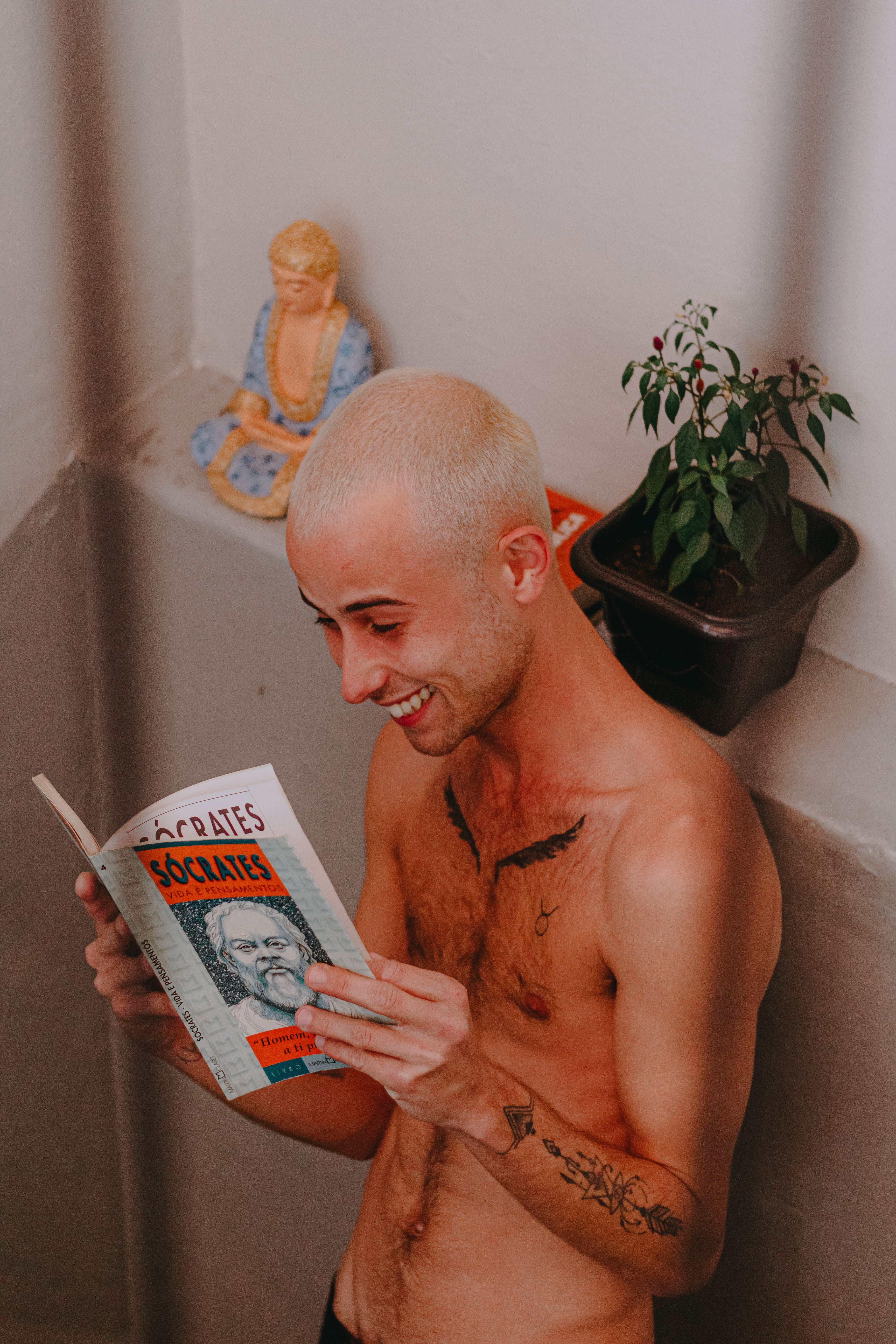 Tattooed man smiling while reading a book indoors with a plant and statue nearby.