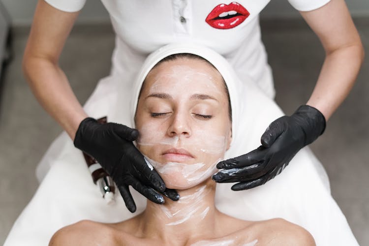 Unrecognizable Beautician Removing Clay Mask From Woman Face