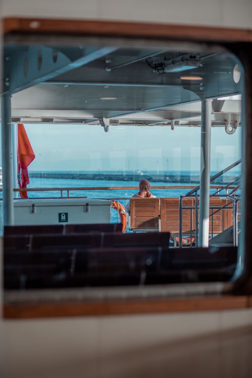 Free A lone woman sits on a ferry, looking out to the sea through a large window, capturing a tranquil moment. Stock Photo