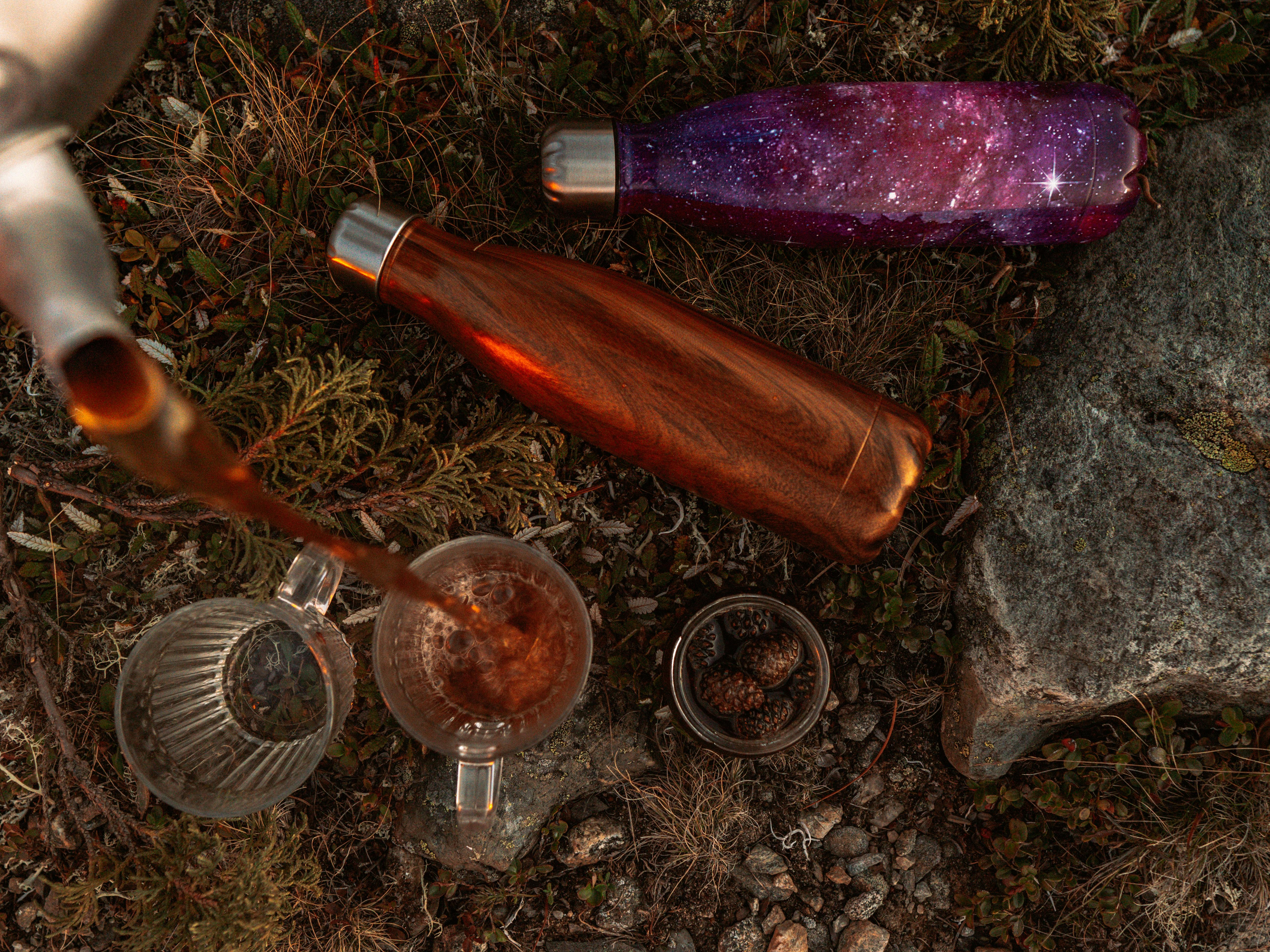 Pouring tea into a glass outdoors with thermos flasks on the ground.