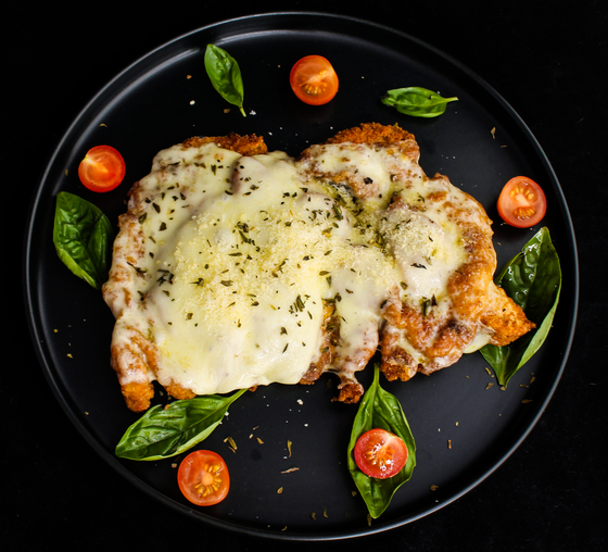 Breaded chicken cutlets covered in melted mozzarella with basil leaves and cherry tomatoes on black plate