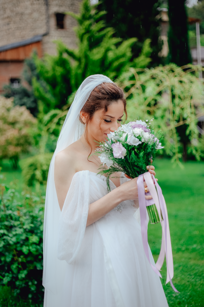 A beautiful bride in a wedding dress gazes at her bouquet in a lush garden setting.
