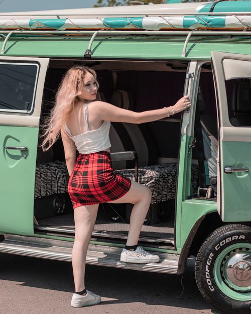 Free A young woman in a checked red skirt poses at the door of a vintage green van on a sunny day. Stock Photo