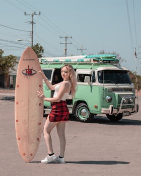 A young woman poses with a surfboard next to a vintage van on a sunny day.