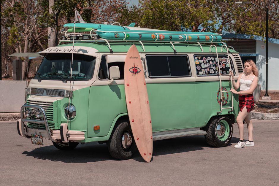 A young woman stands by a retro green van with a surfboard, ready for a summer adventure.