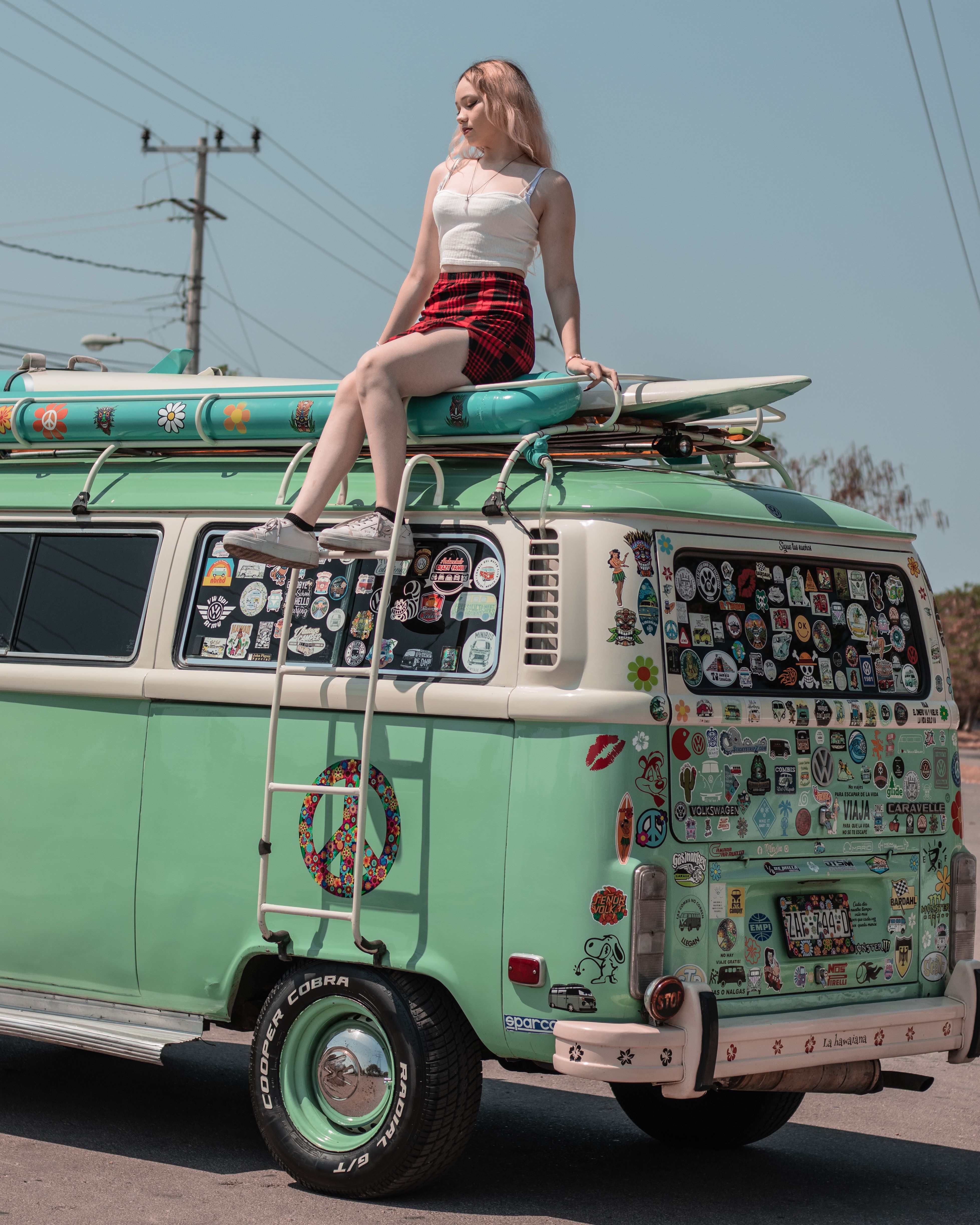 Young woman poses on vintage van decorated with stickers, embodying youthful retro culture.
