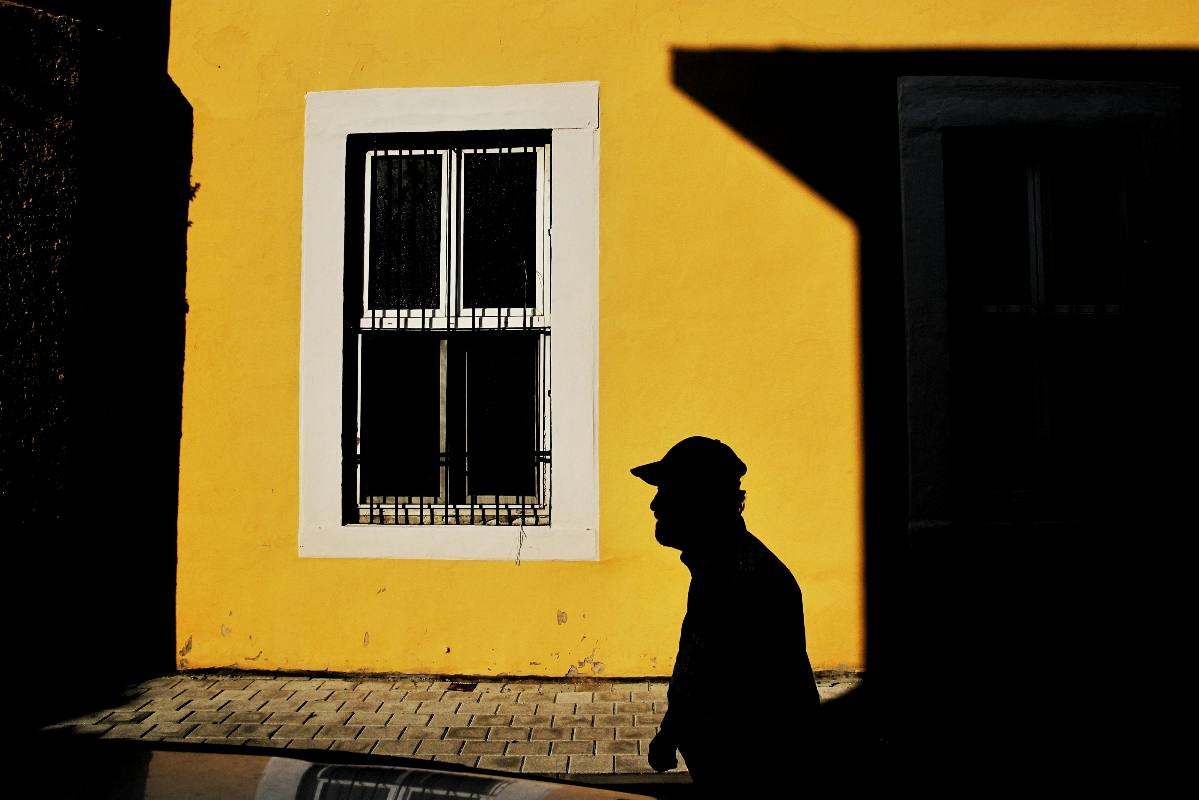 A man in silhouette walks past a yellow wall with window during daylight.