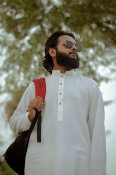 Bearded man in white traditional clothing stands outdoors confidently, holding a backpack.