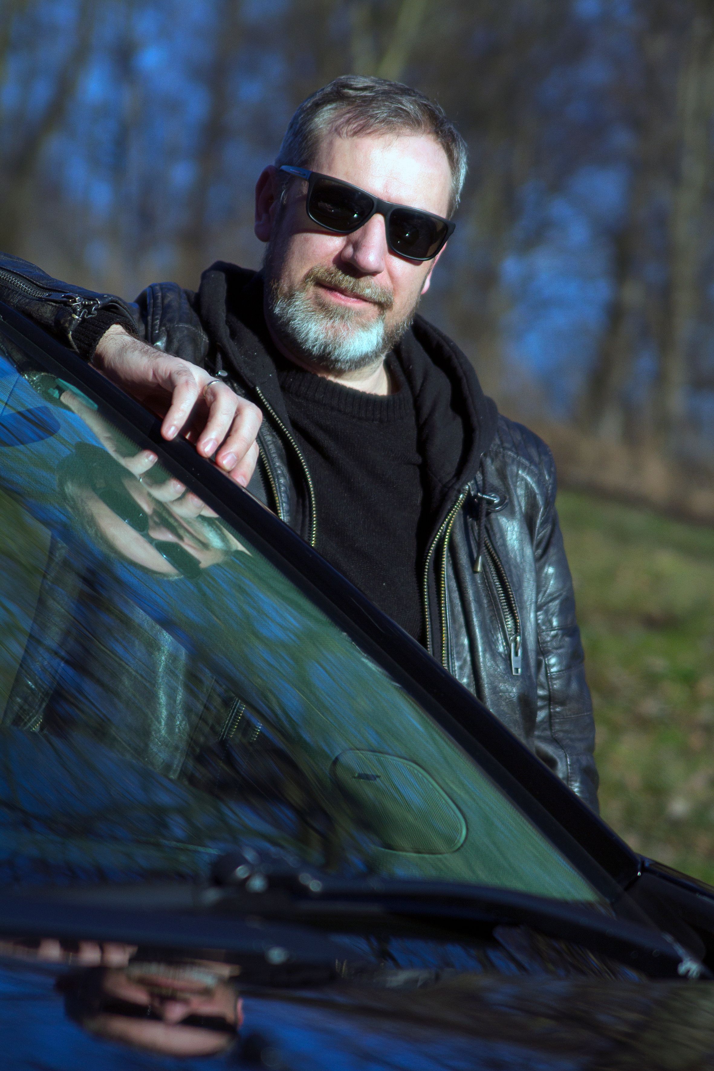 Confident man in sunglasses and leather jacket posing beside a car outdoors.