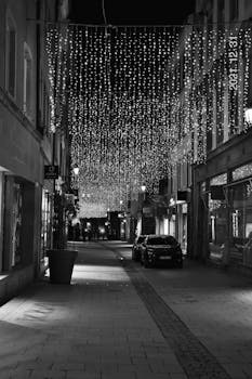 Black and white photo of a European street at night adorned with festive lights and cars.
