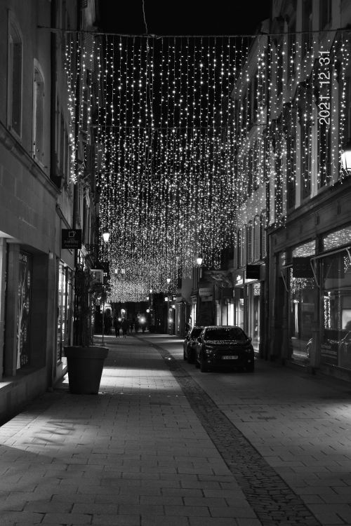 Free Black and white photo of a European street at night adorned with festive lights and cars. Stock Photo