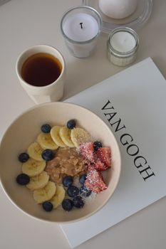 A nutritious breakfast bowl with blueberries, bananas, strawberries, peanut butter, and tea on a Van Gogh book.