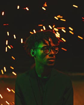 Moody portrait of a young man in glasses, illuminated by fire sparkles at night.