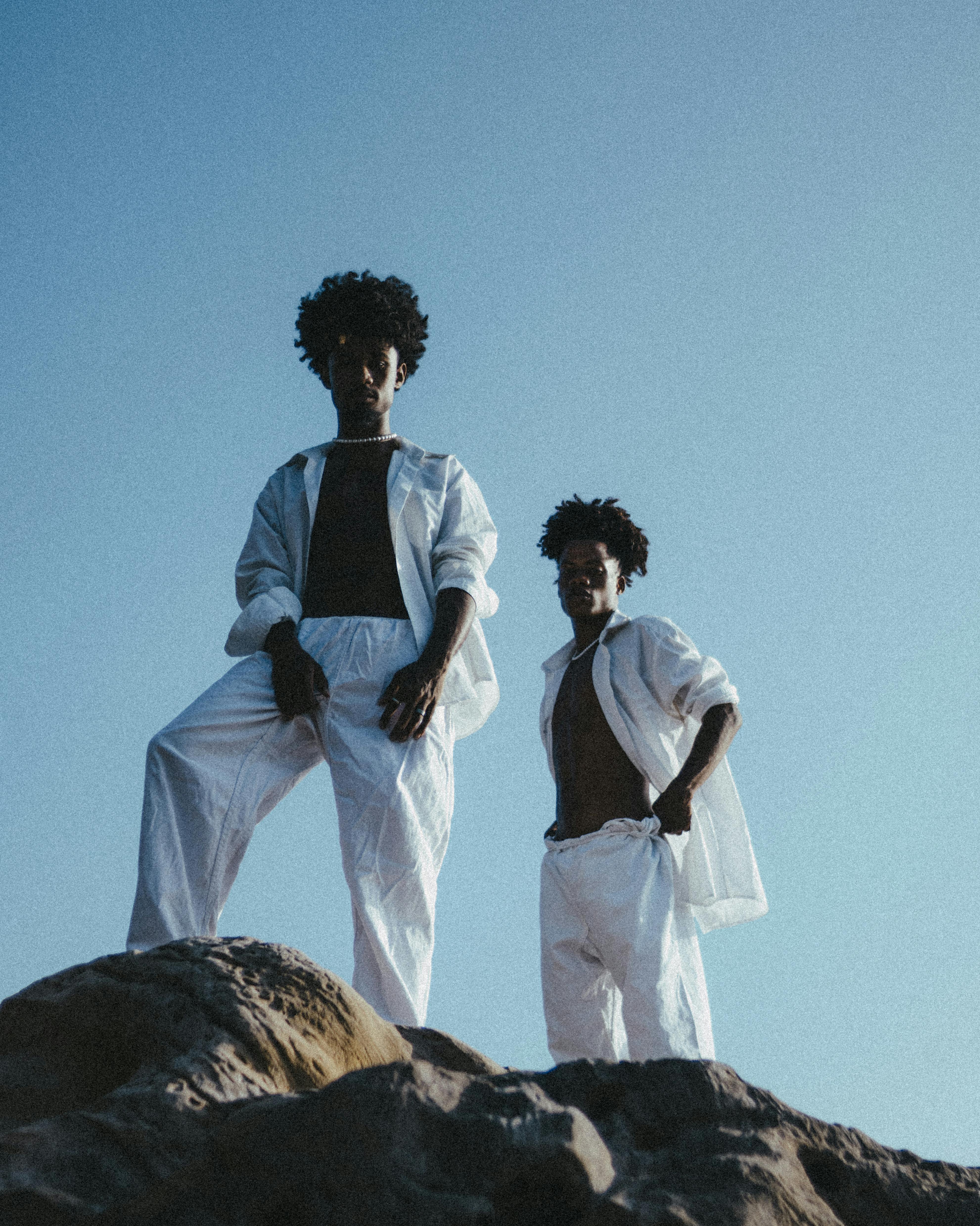 Two young men with Afro hair pose confidently on rocks against a clear blue sky.