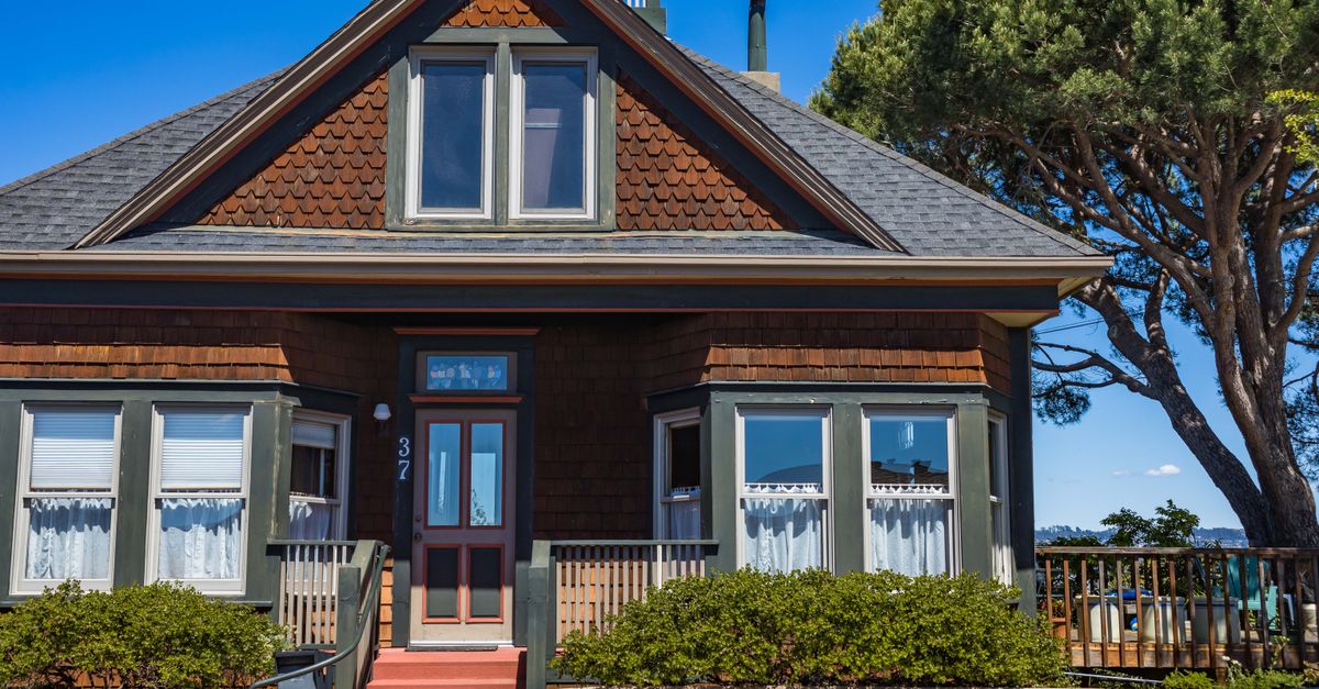 A picturesque vintage house with red stairs, green trims, and bay windows set against a clear blue sky.