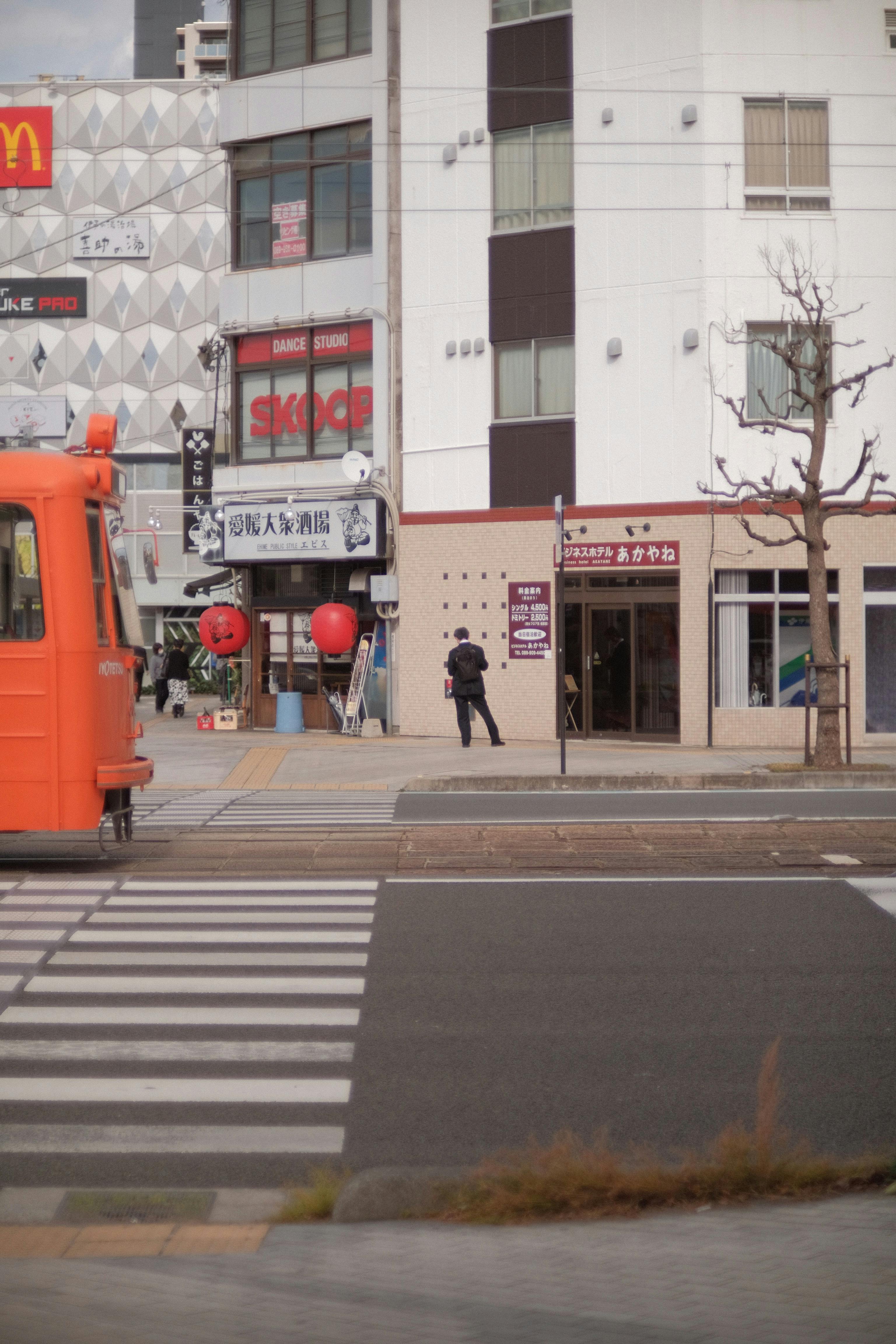 Free A vibrant urban street scene featuring an orange tram and a pedestrian near shops and buildings. Stock Photo