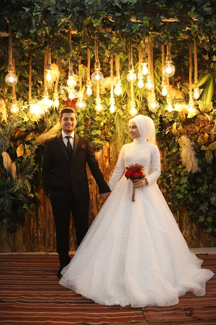 Bride And Groom Standing Under Hanging Lightbulbs And Leaves