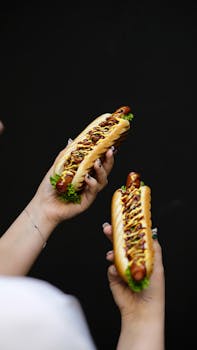 Two hot dogs held against a dark background with mustard, lettuce, and a soft bun.