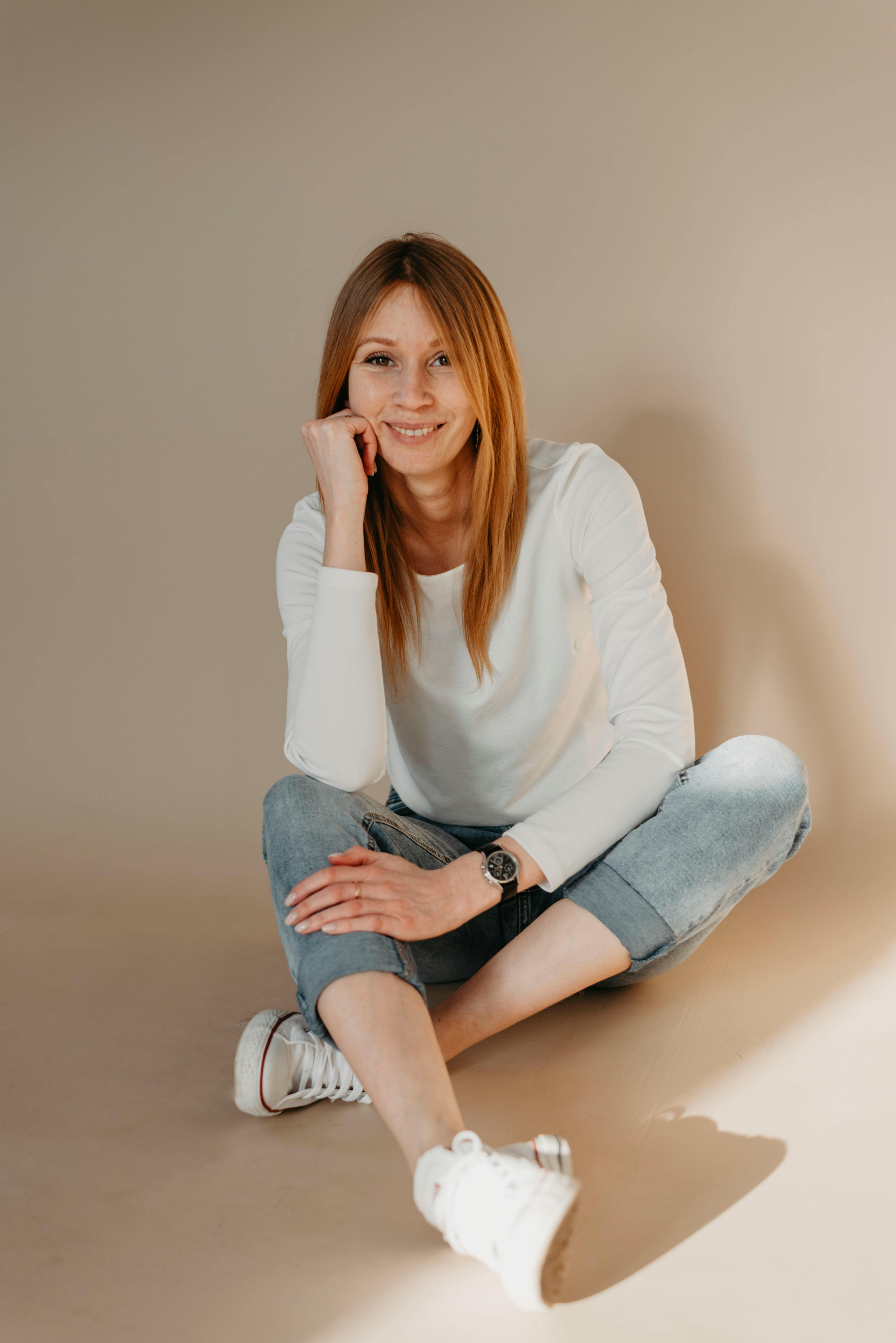 Smiling woman in casual denim and white outfit sitting indoors on neutral background.