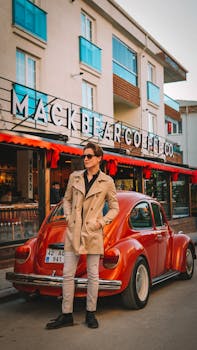 A fashionable man in a beige coat stands by a classic red Volkswagen Beetle on a stylish street.