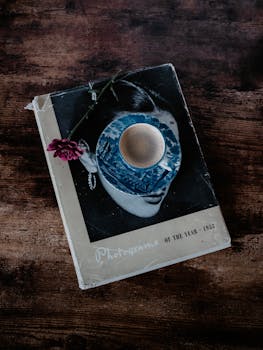 A nostalgic flat lay featuring an old book, coffee, and a single flower on a wooden table.