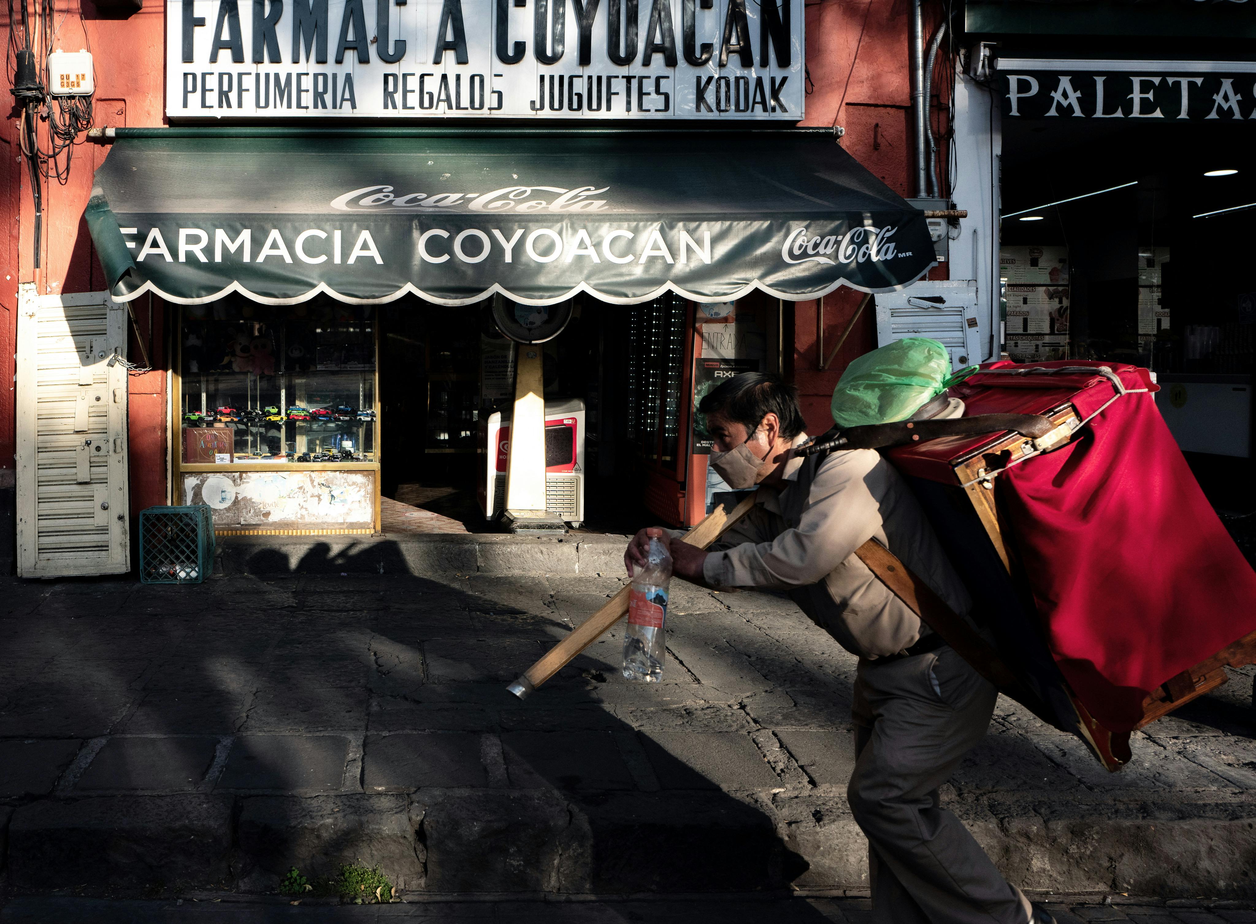 Man carrying goods past a pharmacy in Coyoacán, Mexico City, capturing vibrant urban life.