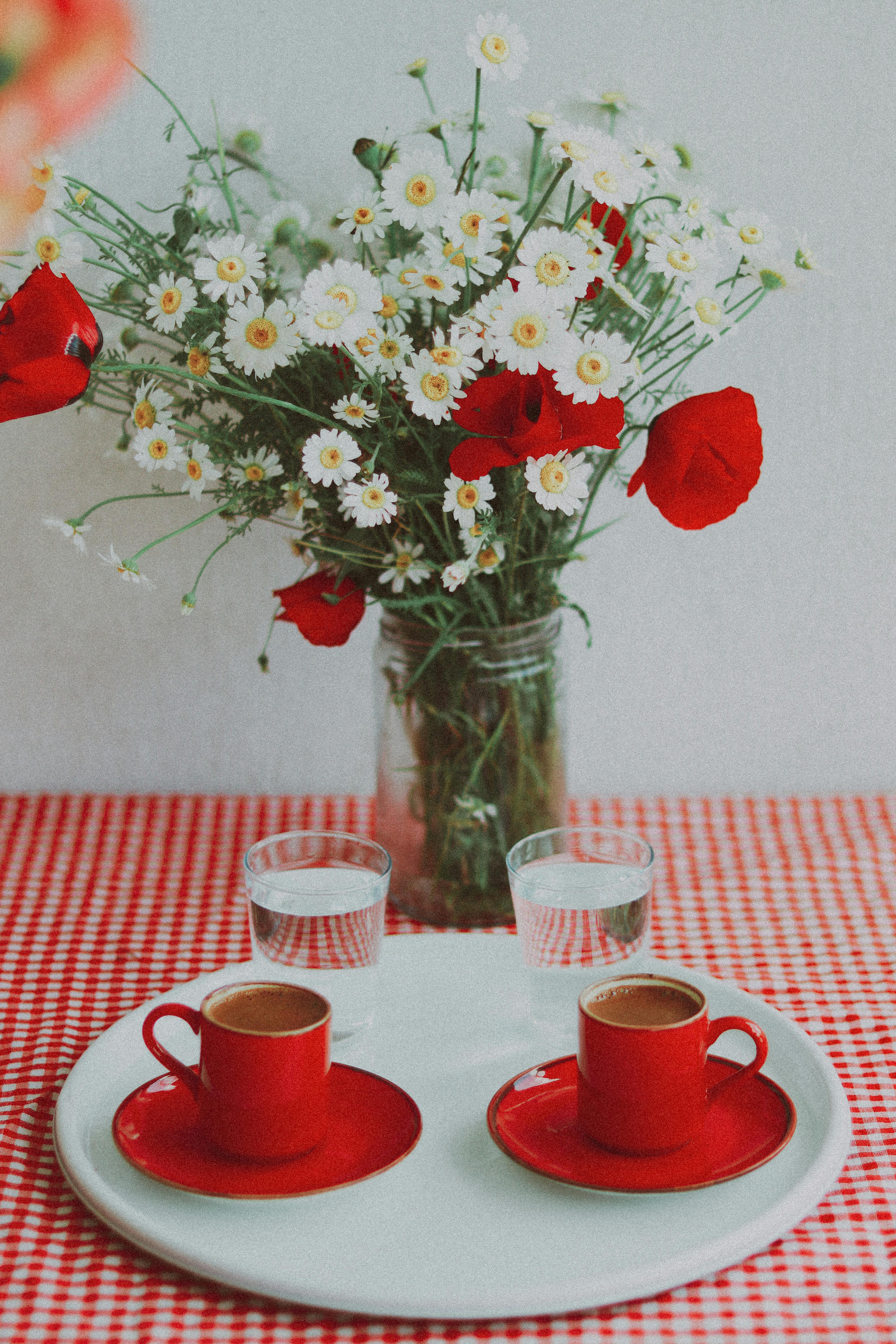 Red coffee cups with saucers on a checkered tablecloth, adorned with a vase of poppies and daisies.