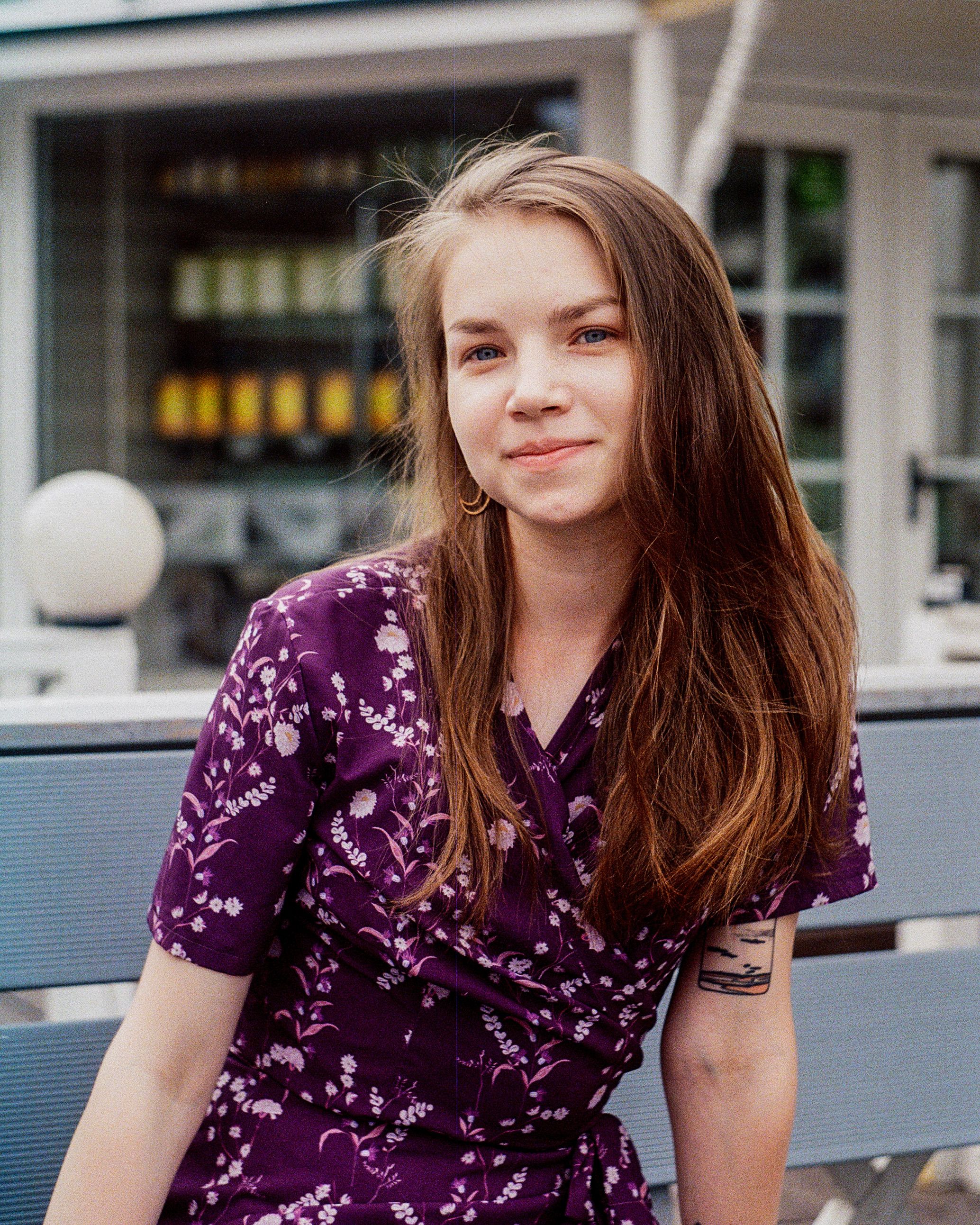 Young woman with long brown hair in a floral purple dress sitting on a bench outdoors.