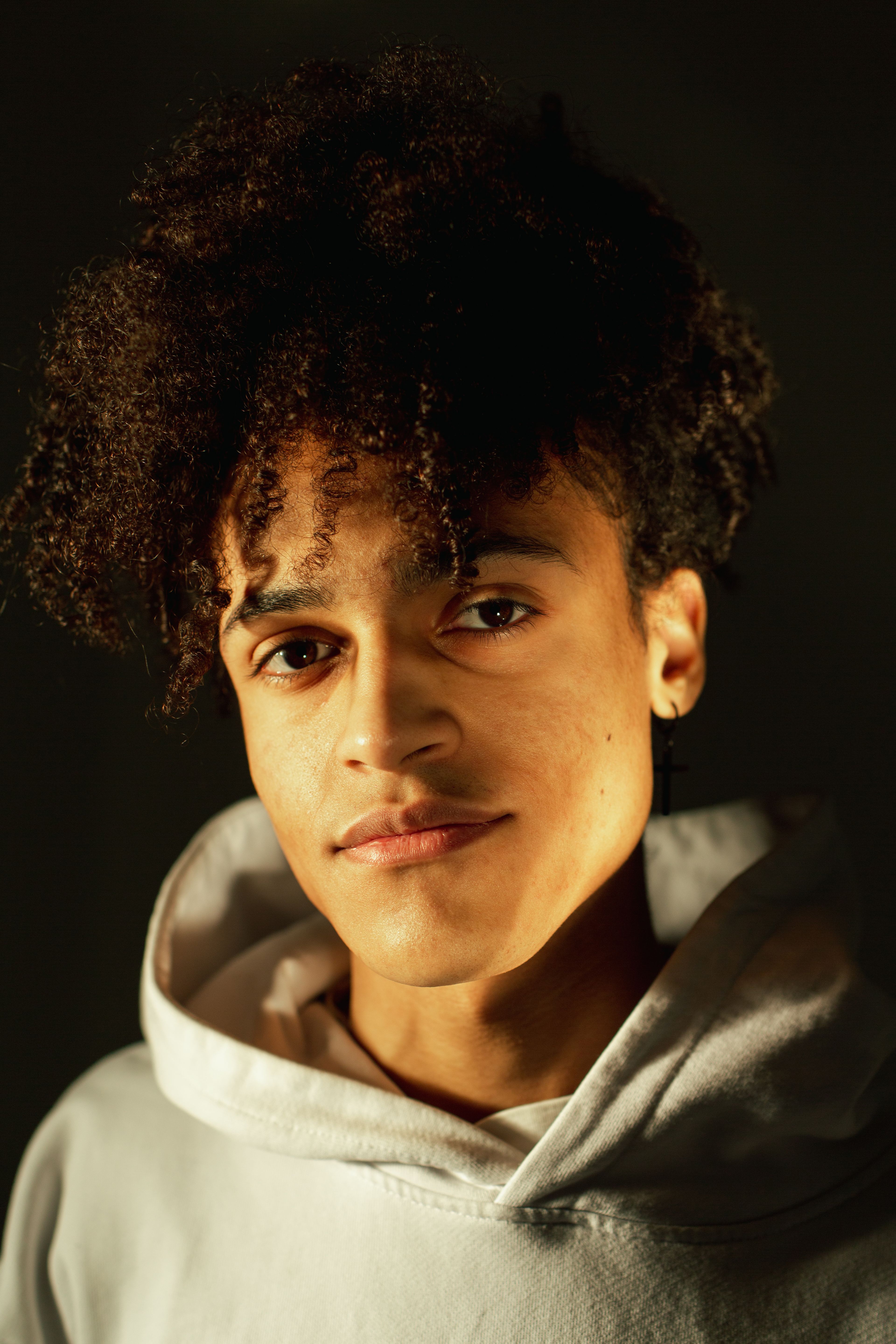 Close-up portrait of a young man with afro hair, wearing a hoodie against a dark backdrop.