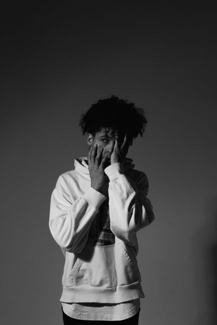 Artistic black and white portrait of a man posing in a hoodie, hands over face.