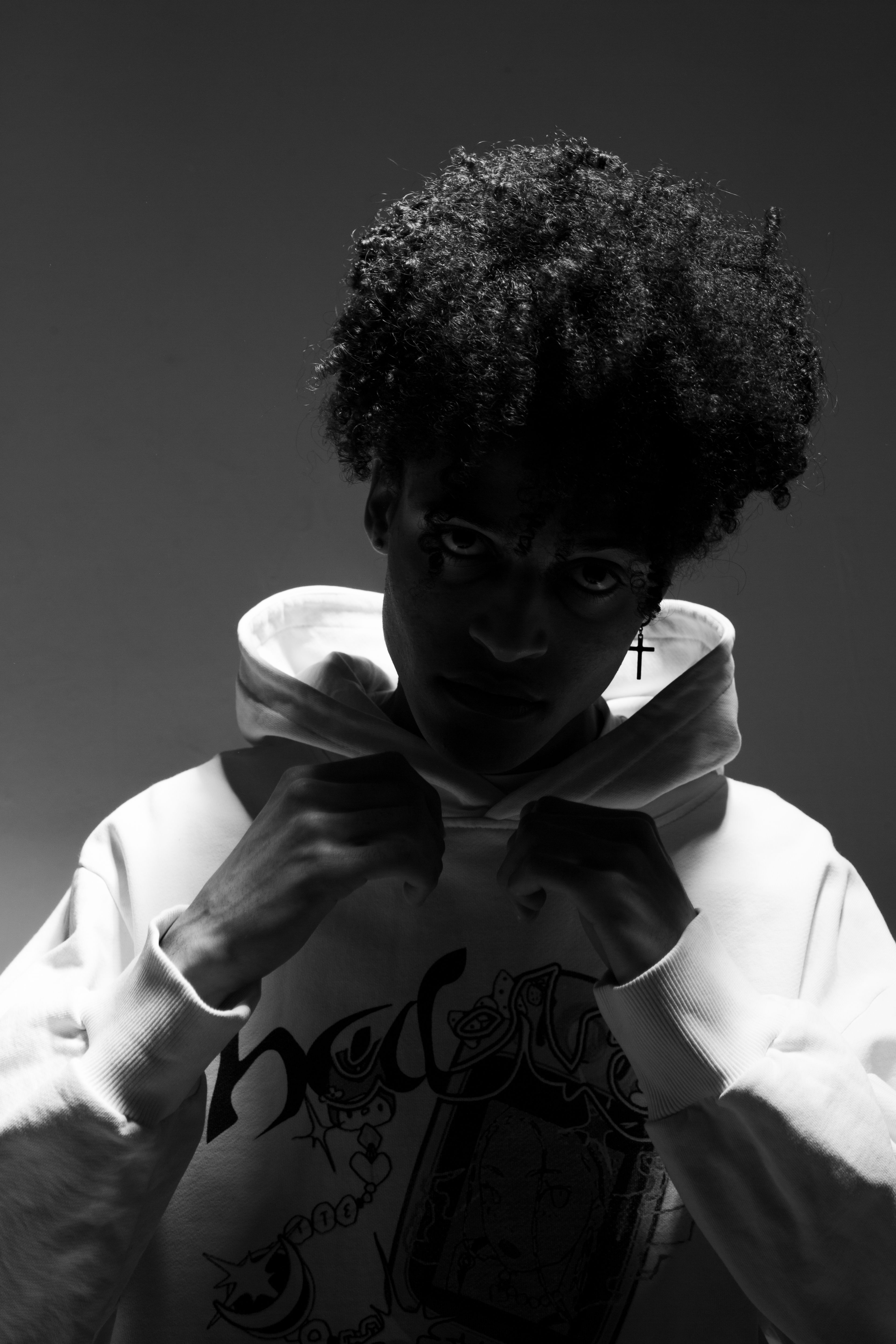 Monochrome portrait of a person adjusting a hoodie, showcasing their afro hairstyle in a studio setting.