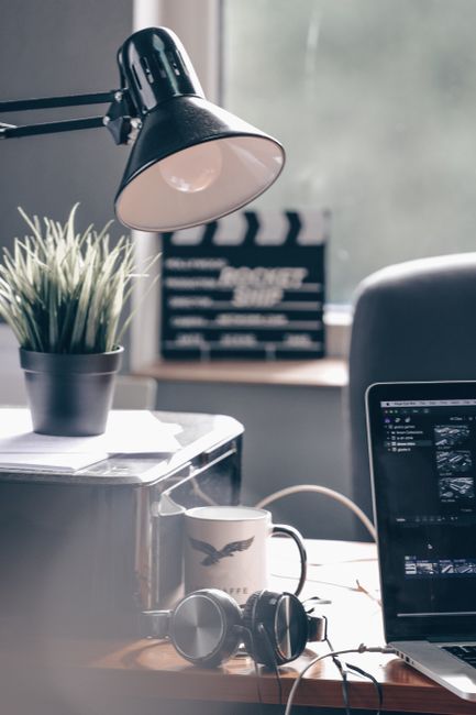 A modern office workspace featuring a laptop, desk lamp, potted plant, and headphones by a window.