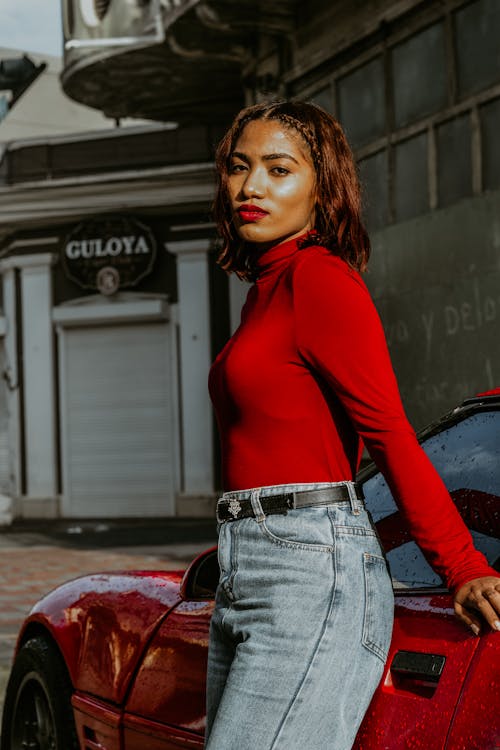 Free Woman in Red Long Sleeve Shirt and Blue Denim Jeans Sitting on Red Car Hood during Stock Photo