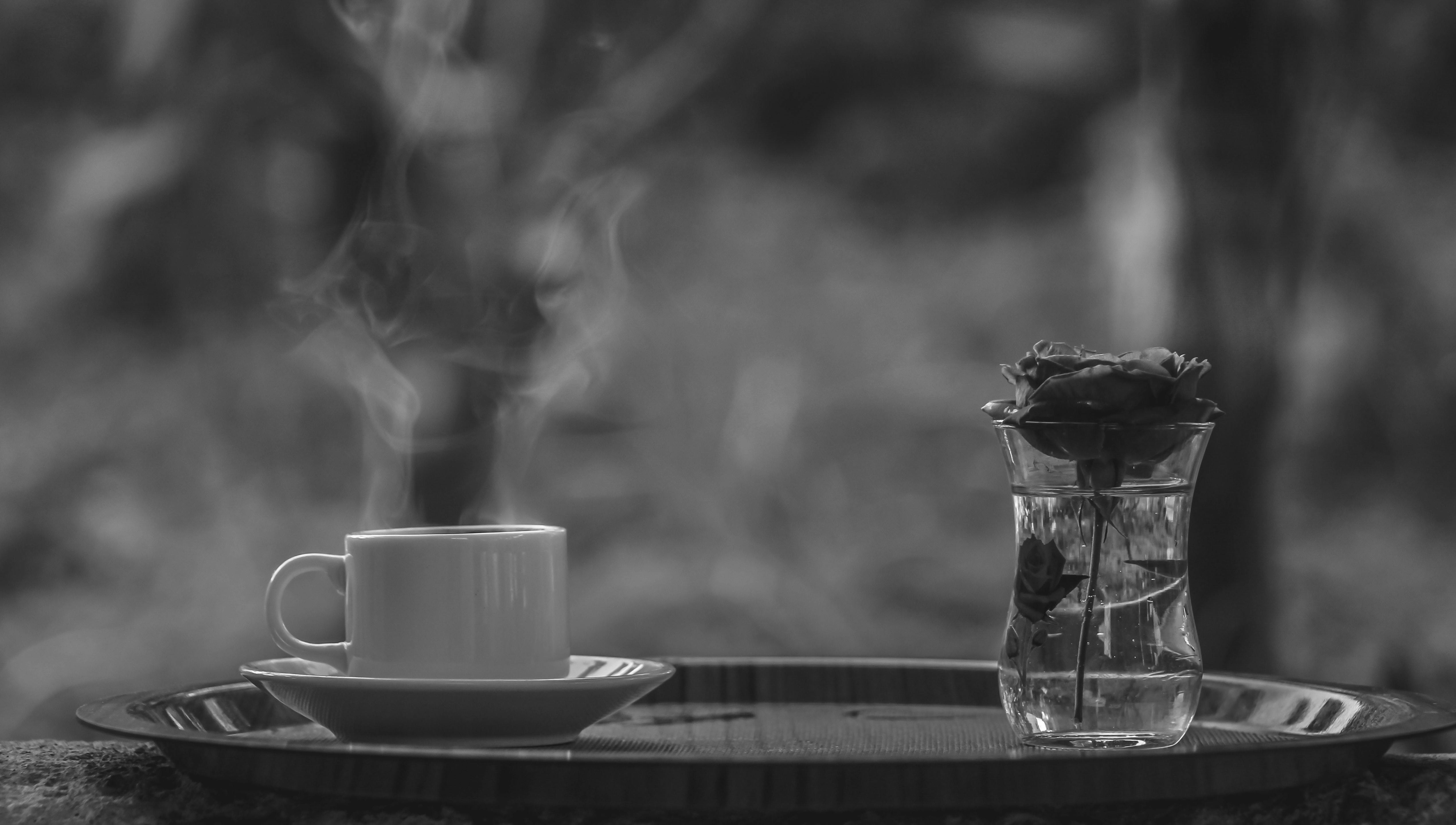 一张单色图像里，有一杯热气腾腾的咖啡，旁边是插在花瓶中的玫瑰