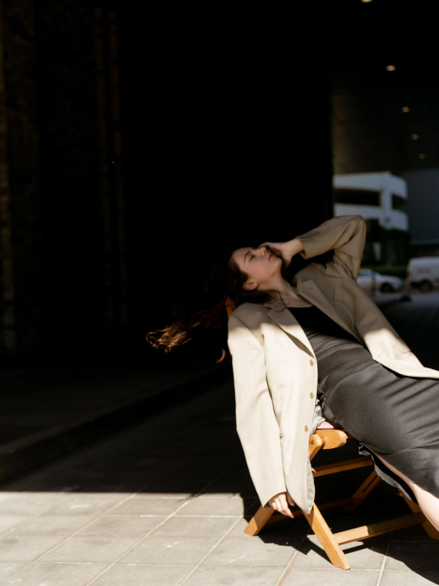 A woman in a brown coat reclining on a wooden chair — soft neutral layering, CBK ease