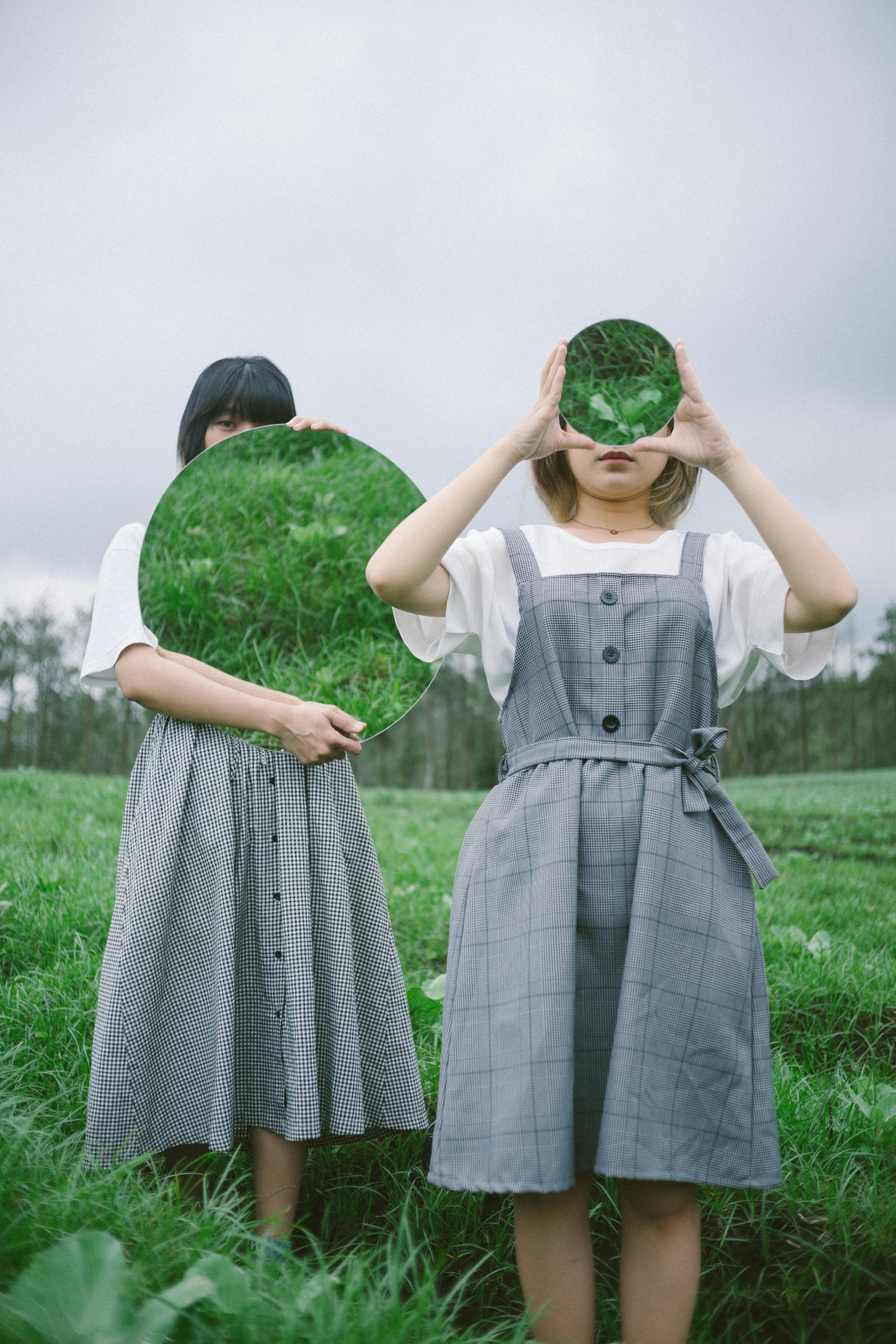 Two women in a grass field holding mirrors, reflecting nature and creating an artistic analogy of identity.