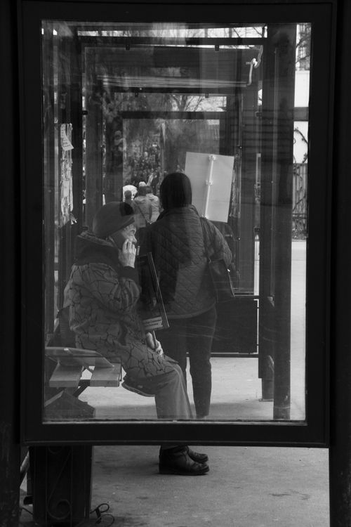 Free Monochrome image of an elderly woman sitting at a bus stop using a smartphone. Stock Photo