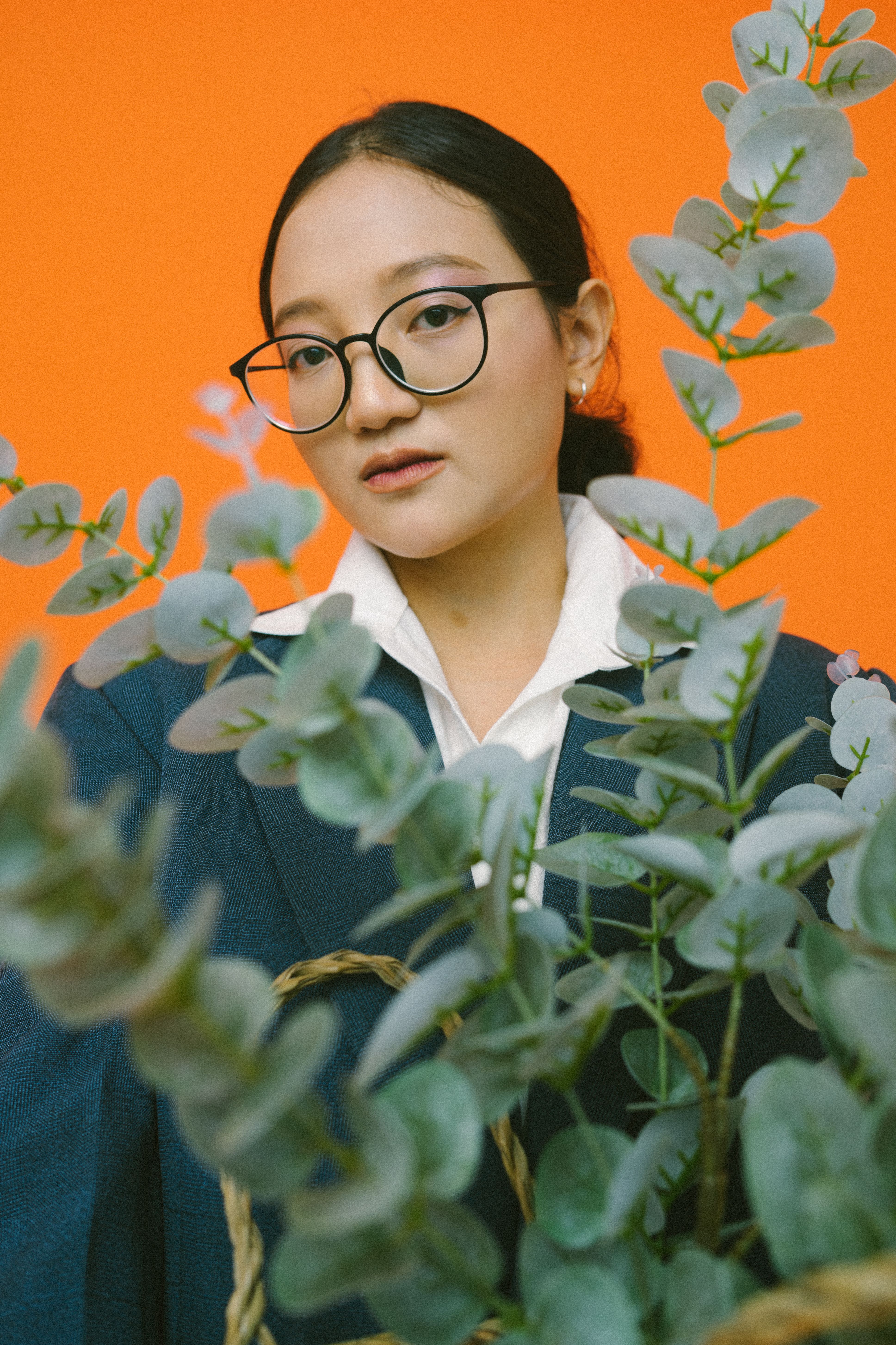 Elegant portrait of an Asian woman with glasses and eucalyptus against an orange background.