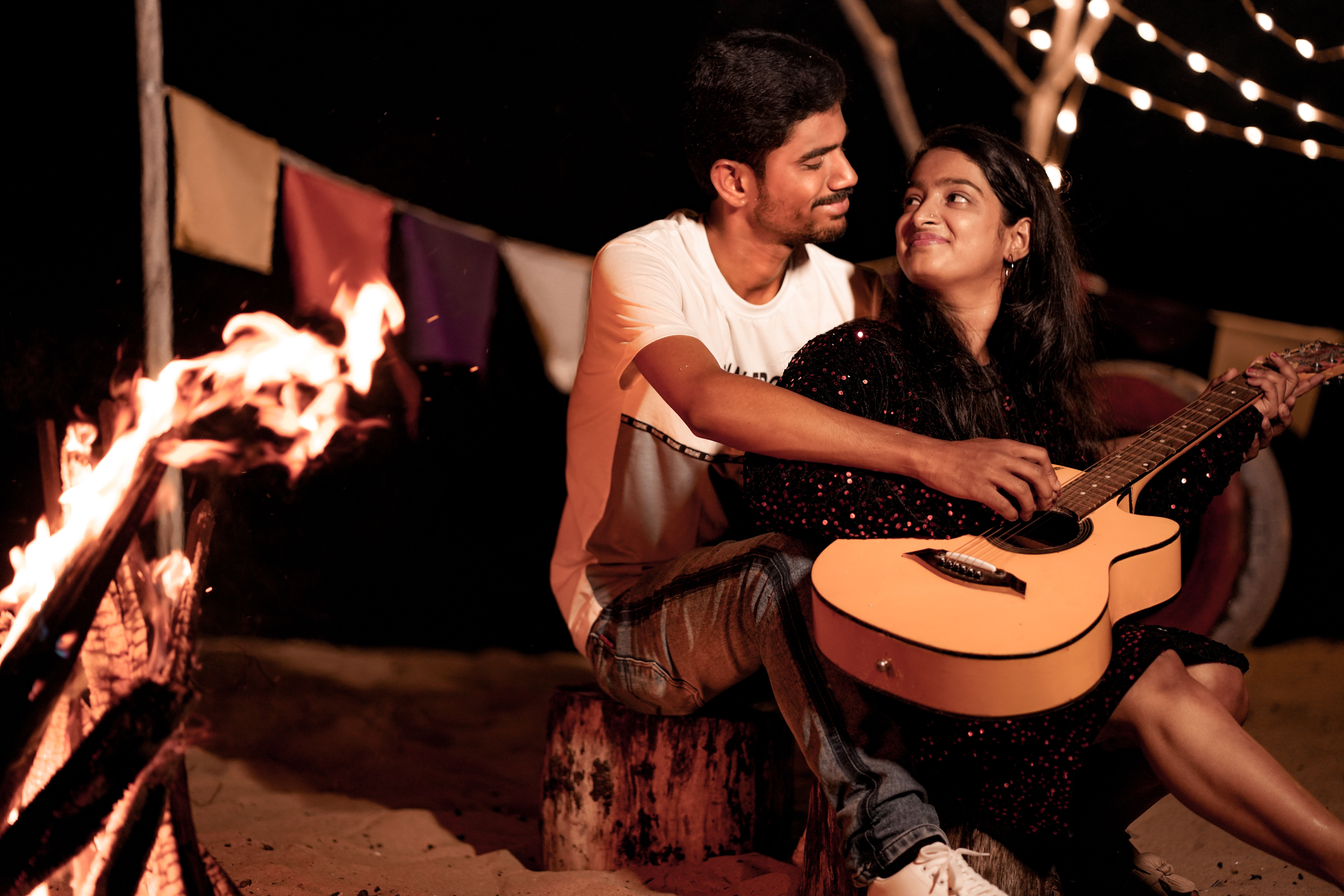 A romantic couple enjoying a night by the campfire, playing guitar under string lights.