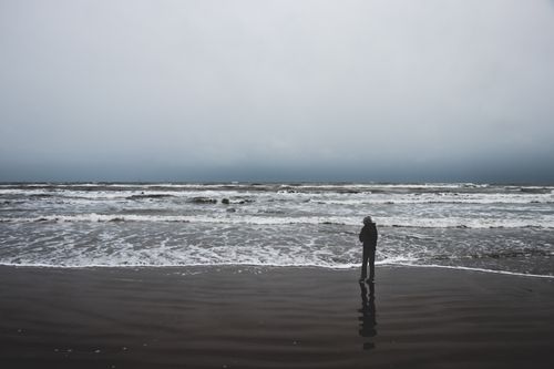 Free A person stands alone on a beach looking at the turbulent sea under cloudy skies. Stock Photo