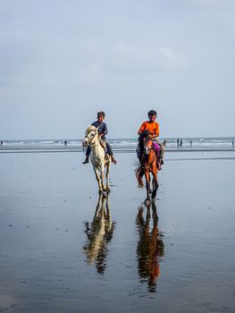 Two men horseback riding on a sandy beach with reflections in the water, showcasing a serene coastal landscape.