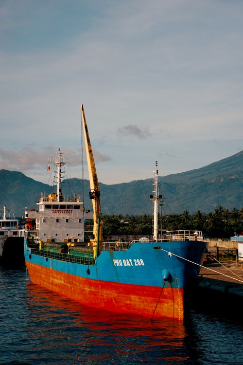 Free A vibrant cargo ship docked at a port with a mountain backdrop, ideal for travel and transportation themes. Stock Photo